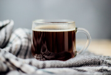 A warm plaid scarf with fringe lies on a light wooden background. A glass mug with black coffee stands on a soft cloth.