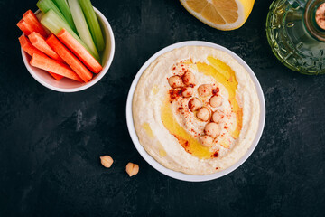 Hummus with olive oil and chickpea in bowl with vegetable sticks on dark stone background.