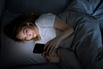 Young woman in bed holding a phone, tired and exhausted, blue light straining her eyes, messing up...