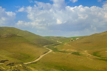 Naklejka premium Country road winding between hills of valley. Village houses and small figures of residents are visible near hill on right side. Shot in Qashqadaryo region of Uzbekistan