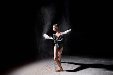 Attractive Caucasian Woman In Black Bodysuit Is Moving Gracefully, In Black Studio With Flour, Athlete Female Is Flexible And Elegant, Make Performance Alone. Art, Dance Concept