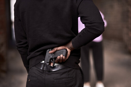 Robbery or criminal man in black hoodie is holding gun standing behind the woman, going to commit a crime. view from back on male in black outfit, close-up hands. criminal activity concept