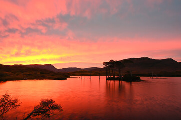 Stunning sunrise over Loch Assynt in Sutherland, Scottish Highlands. On NC500 route. Silhouettes of trees on island on loch, hills in background. Red, orange, yellow and blue sky reflected in water.