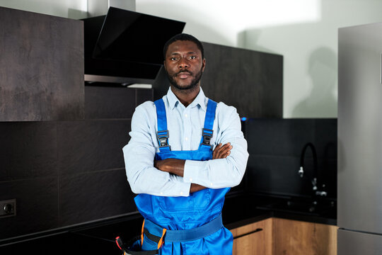 Confident Handsome Young African Handyman Plumber In Blue Uniform. Young Skilled Handyman In Overall Standing At Home In Kitchen Posing Looking At Camera. Repairman Service, Hard Work Concept
