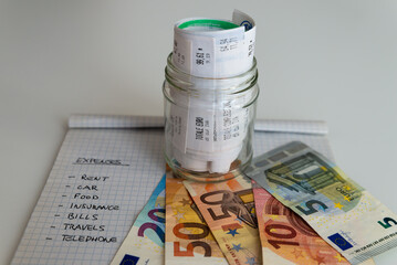 Monthly budget notebook next to banknotes, coins and jar full of rolled up receipts. Increase in tariffs and prices.