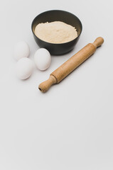 dark stoneware bowl with flour wooden rolling pin and white eggs on a white tabletop