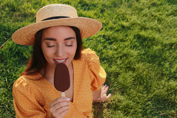 Beautiful young woman eating ice cream glazed in chocolate on green grass outdoors. Space for text
