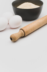 dark gray stoneware bowl with flour wooden rolling pin and white eggs on a white tabletop