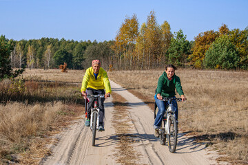 Fototapeta premium Man and woman ride bicycles on a country road through a golden birch grove in autumn
