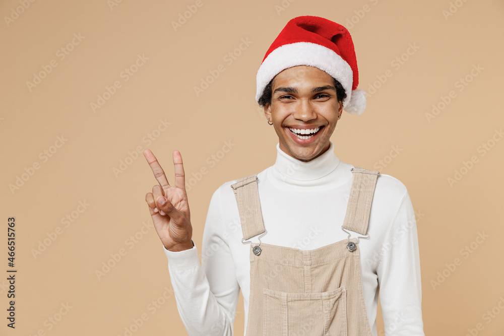 Wall mural Young cheerful smiling happy african man 20s wear Santa Claus red Christmas hat showing victory sign isolated on plain pastel beige background studio portrait. Happy New Year 2022 celebration concept.