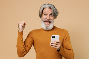 Elderly gray-haired mustache bearded man 50s in mustard yellow turtleneck shirt hold use mobile cell phone do winner gesture isolated on plain pastel beige background studio. People lifestyle concept.