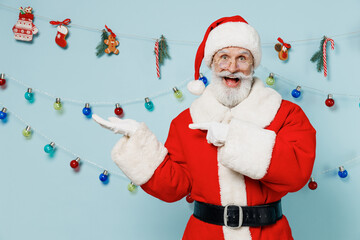 Old Santa Claus man 50s in Christmas hat red suit clothes point finger on empty palm with copy space place mock up isolated on plain blue background studio. Happy New Year 2022 merry ho x-mas concept.
