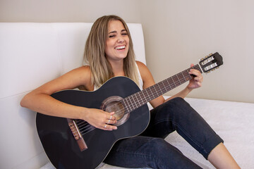 Fototapeta premium Chica sentada en la cama tocando la guitarra. Chica sonriendo. Mujer rubia practicando un hobbie. Disfrutar de la música. Instrumento de cuerda. Guitarra negra.