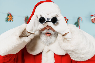Old bearded Santa Claus man 50s wear Christmas hat red suit clothes look in binoculars observe isolated on plain blue background studio. Happy New Year 2022 celebration merry ho x-mas holiday concept