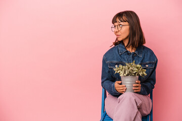 Young mixed race woman sitting on a chair holding a plant isolated on pink background looks aside smiling, cheerful and pleasant.