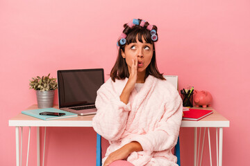 Young mixed race woman in pijama and with curlers isolated on pink background  is saying a secret hot braking news and looking aside
