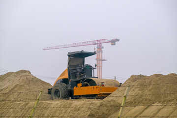 Yellow road compactor on a construction site. Construction equipment on the background of an object under construction. Tower cranes on a construction site.