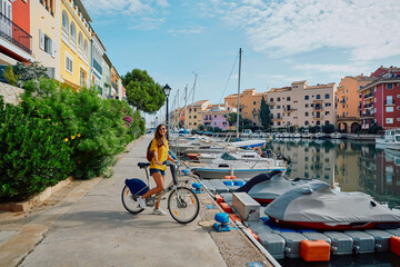 Smiling woman tourist with bicycle on pier with yachts and boats at beautiful harbor of Port...