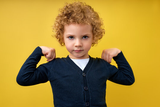 Look at my muscle, I'm strong. Portrait of adorable little boy in T-shirt feeling powerful and self-confident, showing strength. indoor studio shot isolated on yellow studio background