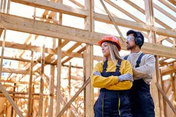 Naklejka na ściany i meble team of constructor engineers in New wooden residential construction home framing. Construction site. Beautiful caucasian lady and man in workwear, hardhat and headset looking at side, at sunny day