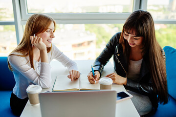 Cheerful diverse female students with modern netbook and education textbook for collaborative e learning discussing web publication in coworking space, happy women enjoying brainstorming studying