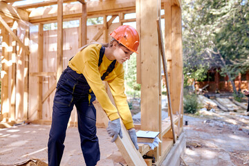 Hardworking caucasian female working on wooden construction site, wearing working uniform clothes, in gloves and hardhat, replacing wooden beam into another place, alone, outdoors.