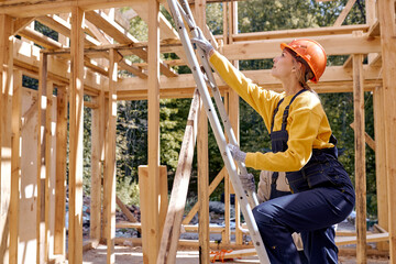 Fototapeta na wymiar Ideas for renovating and extending wooden houses. Female on ladder,goes up to the roof. Hardworking Caucasian Female in working uniform and safety hardhat working on unfinished wooden building site.