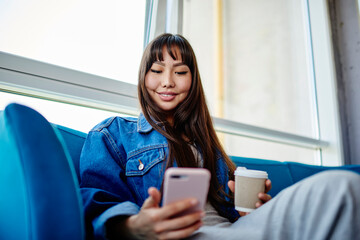Happy Asian hipster girl with takeaway caffeine beverage enjoying positive network messaging in social media, cheerful female blogger with coffee to go reading received email on smartphone device