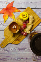 Autumnal still life - bunch of grapes, ripe quince, ginger root lie on wooden cutting board top view. Orange maple leaf on table. Yellow cup with hot aromatic tea drink decorated with flower.