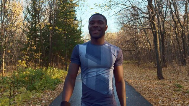 A smiling African American walks slowly down the road in a city prak. Trees without leaves on the branches and evergreen spruces in the background. Autumn, fallen leaves. High quality. 4k footage.
