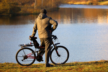 Fototapeta na wymiar Man stopped with bicycle on lake and autumn forest background. Cycling, travel and leisure in fall season