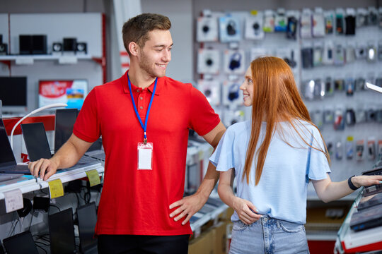 woman is looking for the best laptop in computer department of electronics store. Smiling buyer chooses laptop in modern technology store, pleasant male consultant help customer, have conversation