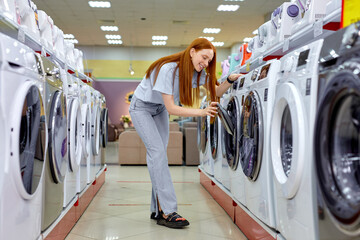 Woman buying new clothes washer in supermarket, choosing the best and the most modern. Redhead Lady In Casual Wear Standing Between Various Different Washine Machines, Make Choice and purchase