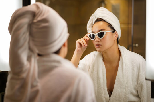 Asian young woman in a bathrobe and hair wrapped in towel looking at the mirror in the bathroom