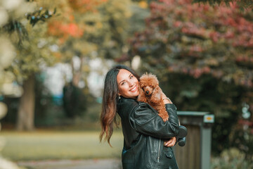 A beautiful young dark-haired woman walks in the park and hugs her poodle dog. Autumn mood.