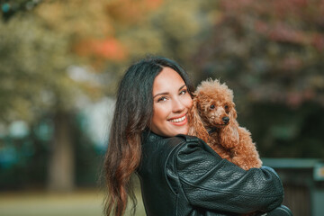 A beautiful young dark-haired woman walks in the park and hugs her poodle dog. Autumn mood.