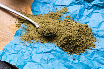Big pile of green powdered kratom on blue crampled paper with teaspoon around