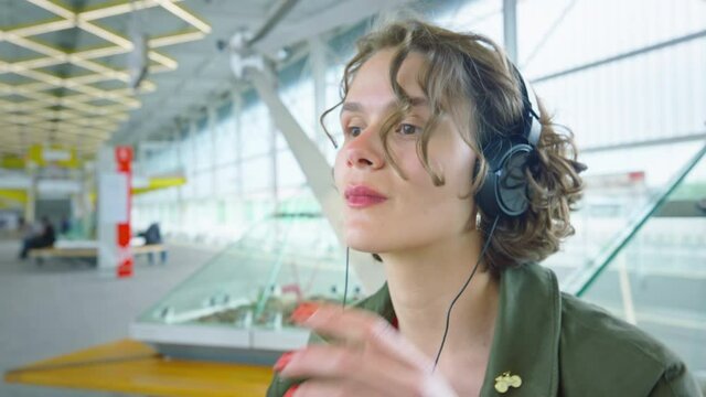 Side portrait shot of young authentic millennial woman sit on bench at train station in arrival or departure hall, put on headphone to listen to audio podcast or music from streaming platform