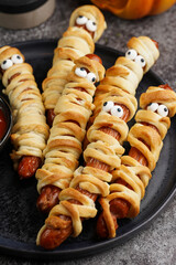 Cute Halloween party snack: wiener sausage with sugar eyes wrapped in dough stripes resembling mummies on a black plate with tomato ketchup