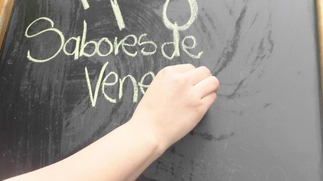 Close-Up of a Female Hand Writing "A Venezuelan Spot in Medellin" with Chalk on a Blackboard