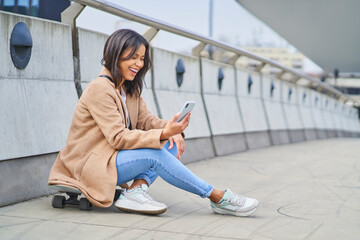 Laughing woman using smartphone sitting on skateboard in the city