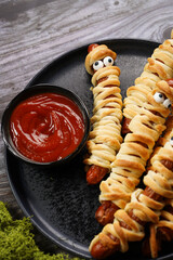 Cute Halloween party snack: wiener sausage with sugar eyes wrapped in dough stripes resembling mummies on a black plate with tomato ketchup