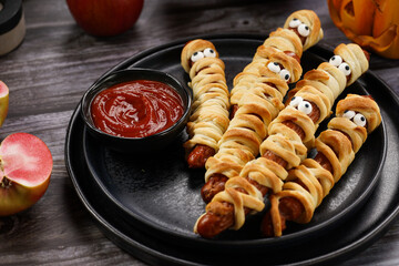 Cute Halloween party snack: wiener sausage with sugar eyes wrapped in dough stripes resembling mummies on a black plate with tomato ketchup