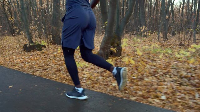 Jogging in a city park against the background of yellow fallen leaves after rain. Development of sports skills. A dark-skinned athlete runs down the road, a side view. High quality. 4k footage.