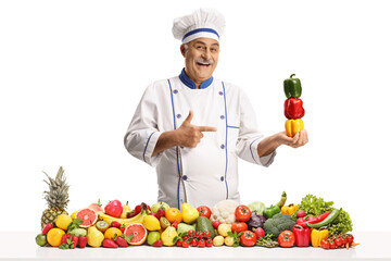 Mature male chef holding bell peppers behind a pile of fruits and vegetables and pointing