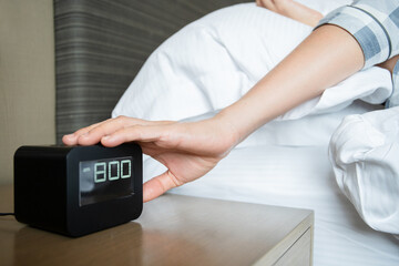 Lazy Asian young woman trying to snooze awaking digital alarm clock on the table beside the bed.
