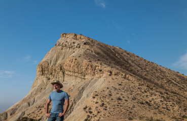 Fototapeta na wymiar Portrait of adult man in cowboy hat in desert area against mountain and blue sky