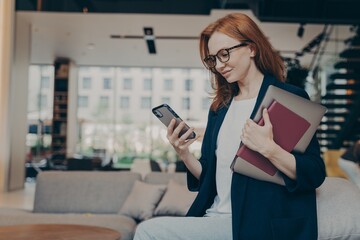 Young smiling businesswoman in glasses with laptop and agenda under her arm looking at smartphone