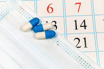 Three blue and white capsules lie on a surgical mask at an angle on a calendar with black and red dates. COVID-19 concept.
