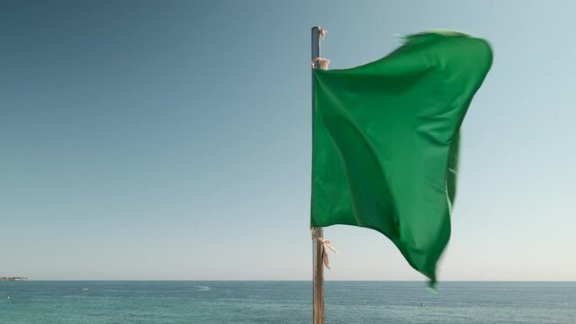 Close up green flag flies against blue sky and Mediterranean Sea background during sunny summer day. Symbol of award given to bathing beach that meets EU standards of cleanliness. Costa Blanca. Spain